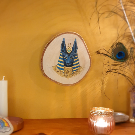 Illustration d'Anubis avec de la pyrogravure et de la peinture sur une tranche de bois de bouleau. Les couelurs utilisées sont dans les bleus foncé et indigo, ainsi que du doré.